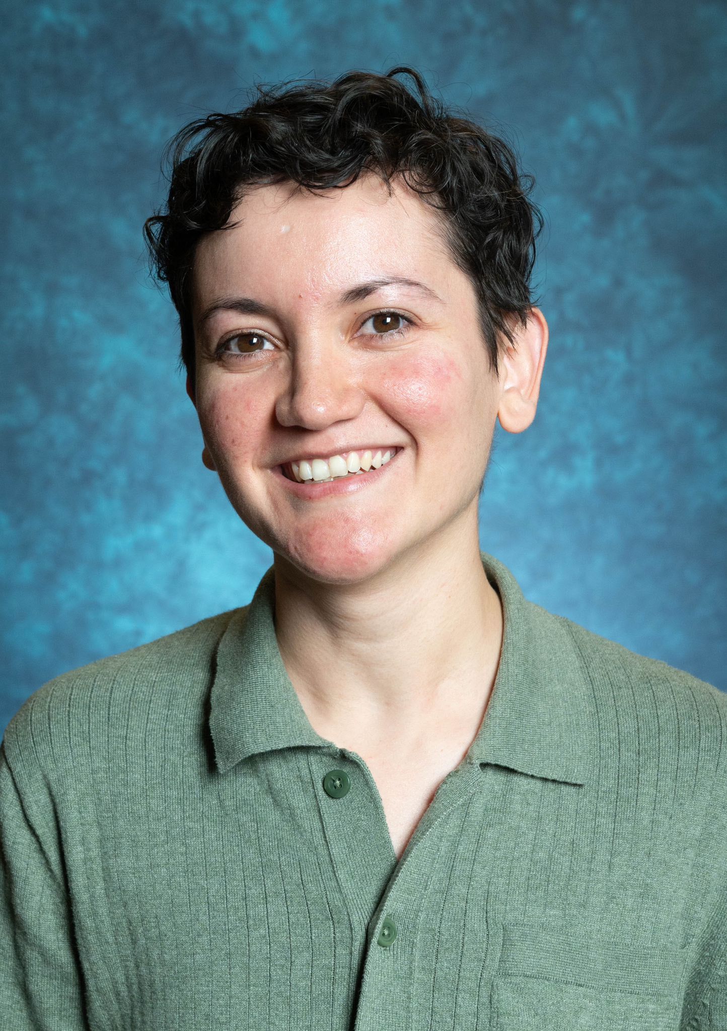 Image of Dr. Rio Lopez, Director of the Latin American Program. The image portrays a person with short, dark, curly hair and smiling directly at the camera. The person is wearing a sage green, collared knit sweater with a ribbed texture and a pocket on the right side. The sweater has button details with one button visibly fastened. The background is an abstract blue, with varying shades creating a textured appearance. The person’s skin has a natural complexion with some visible redness on the cheeks.